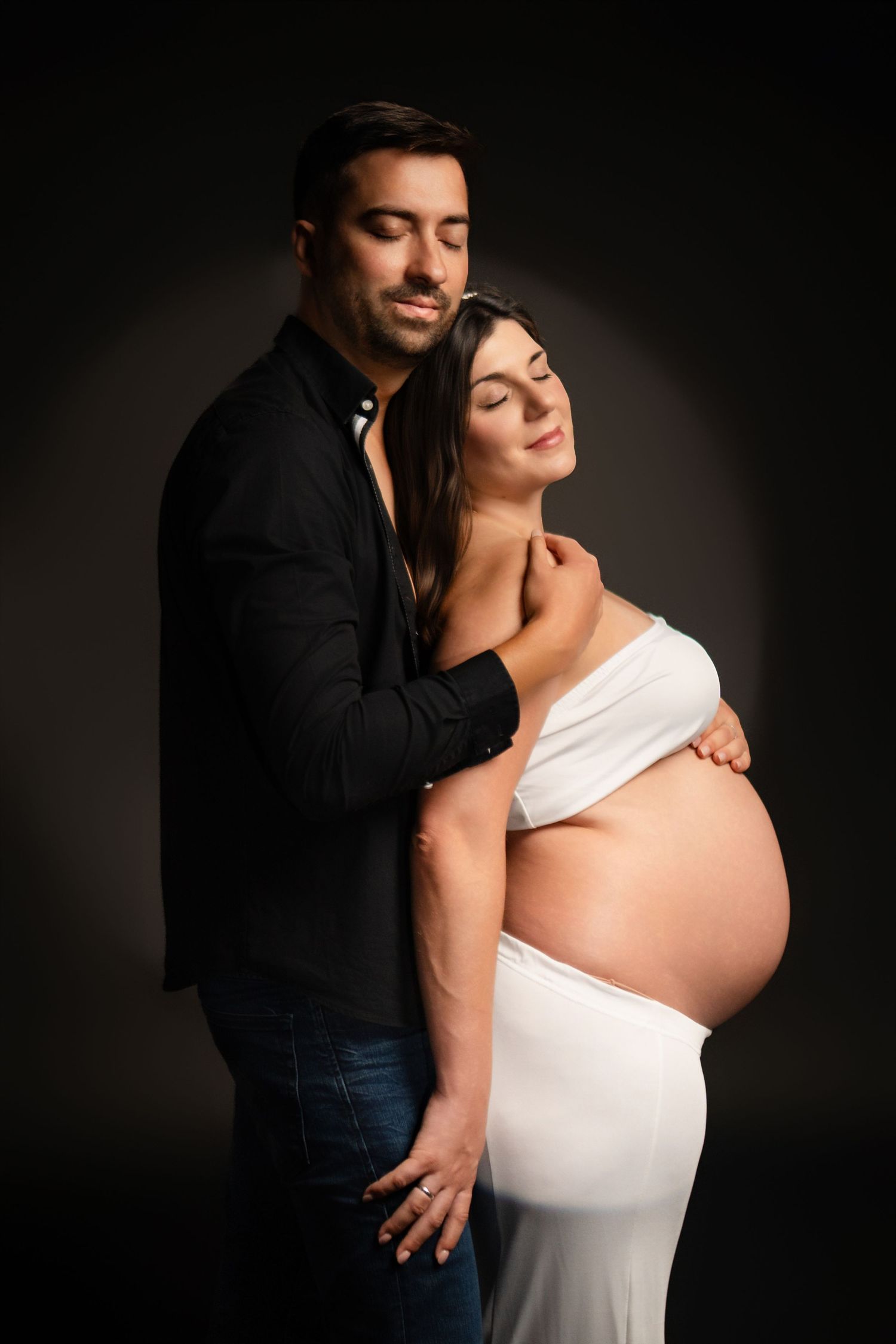 Intimate studio maternity session with dramatic lighting and white flowing gown.