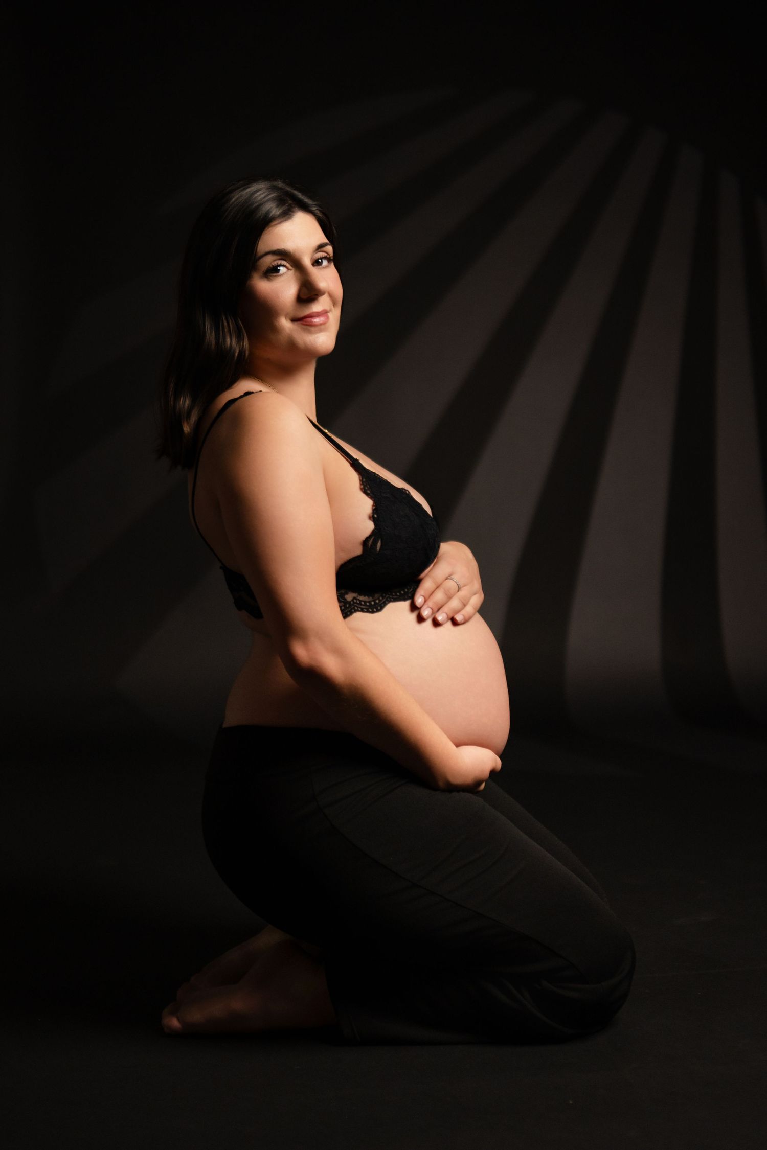 Artistic maternity photo of an expectant mother in black against dark background with dramatic lighting.