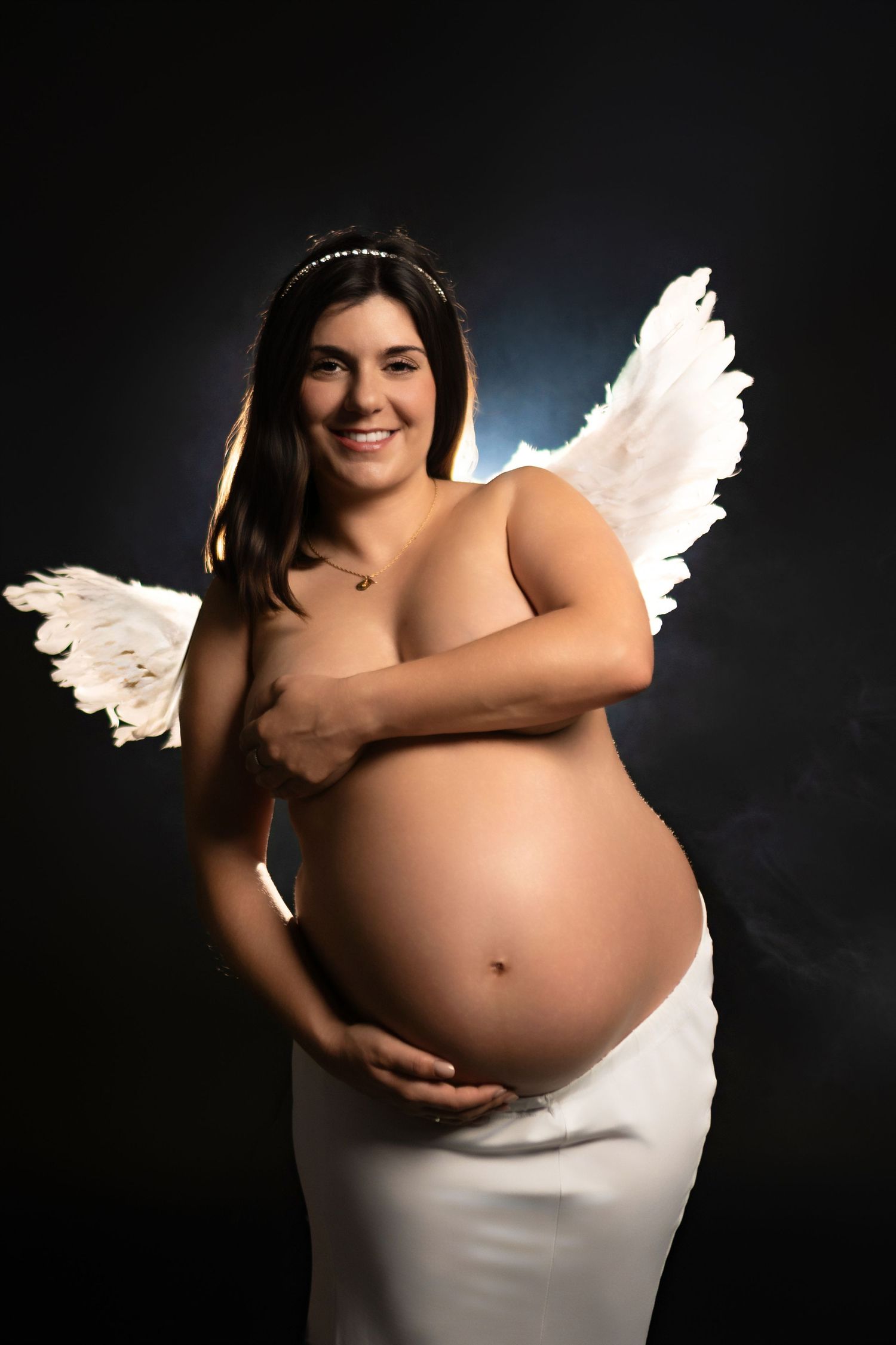 Ethereal maternity portrait featuring white angel wings and dramatic lighting against dark background.