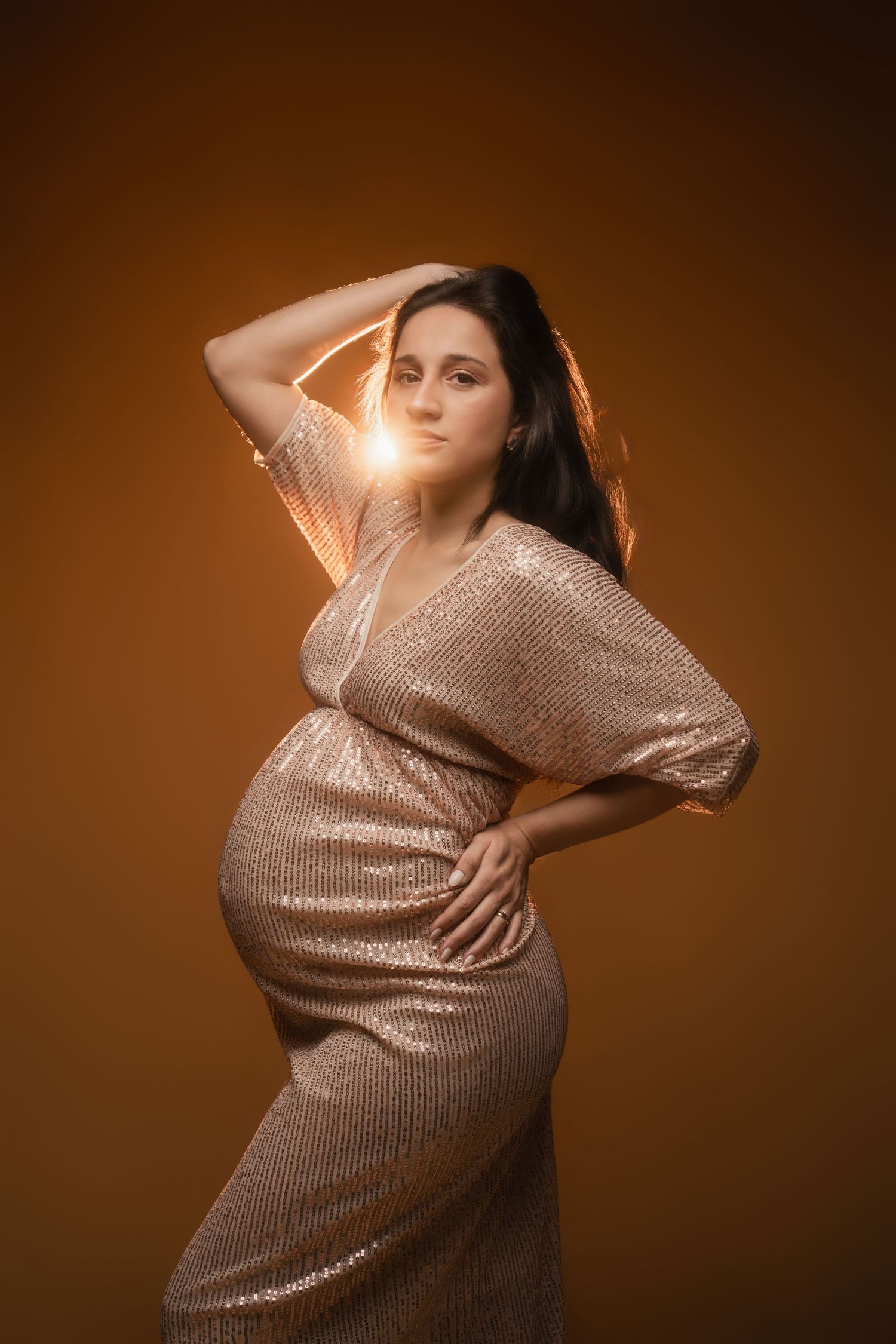 Expectant mother poses in shimmering gold sequin dress against warm brown backdrop in elegant maternity photoshoot.