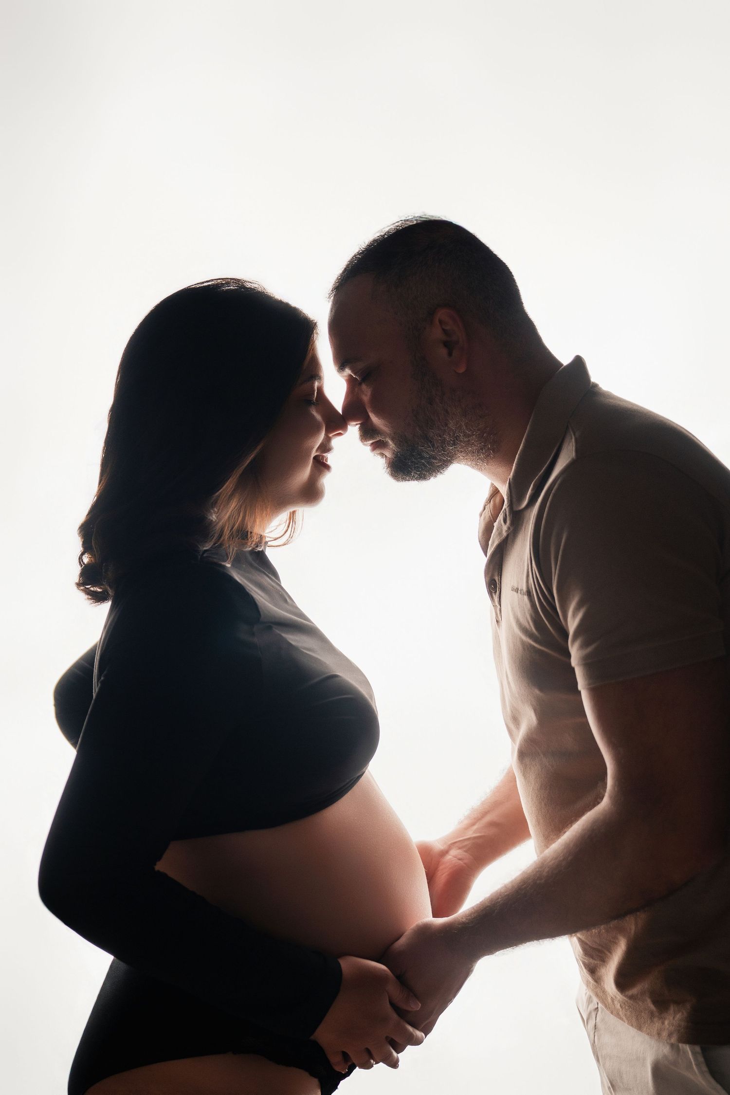 Silhouette of pregnant couple holding hands and touching foreheads in romantic backlit studio portrait.