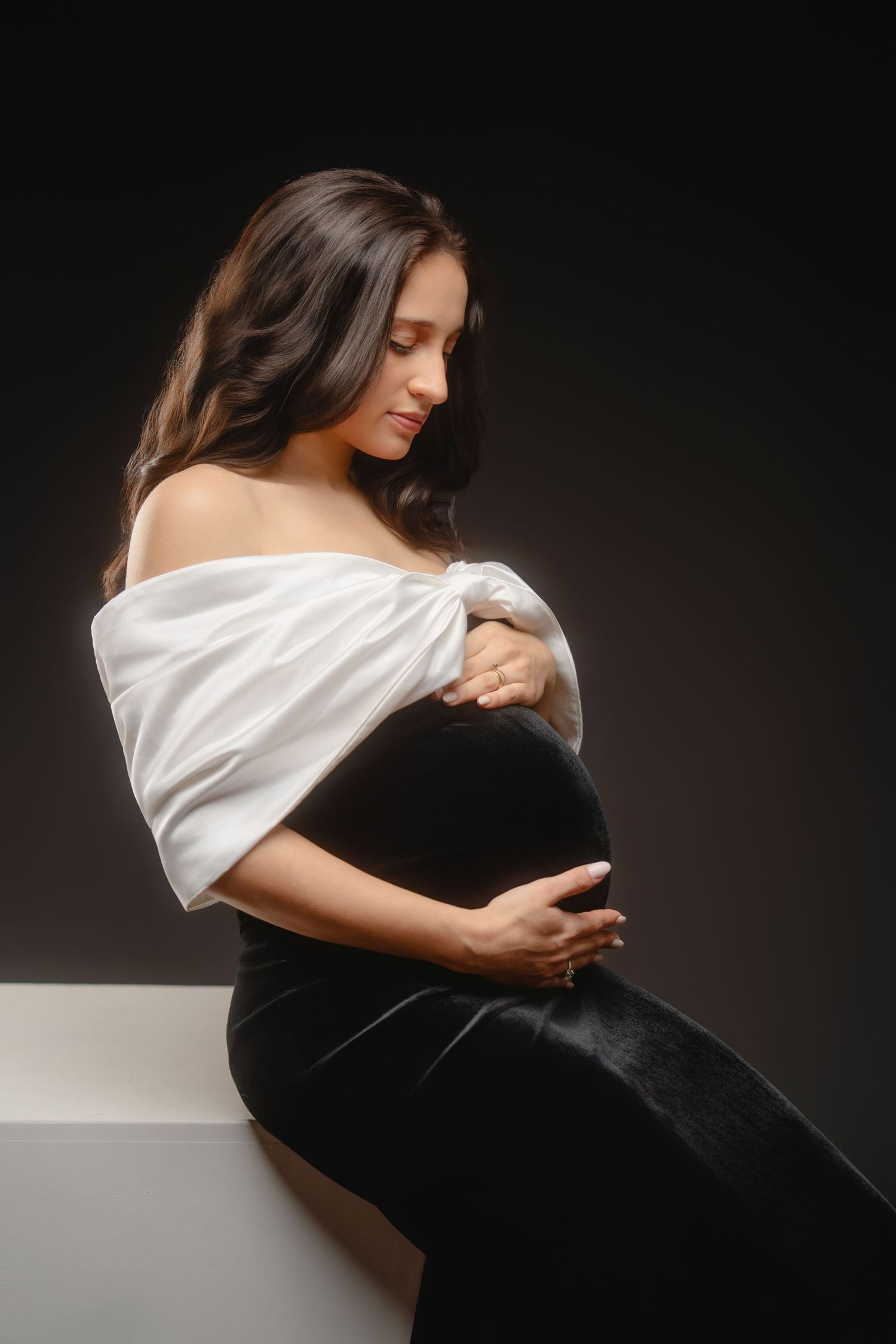 Artistic maternity photo with off-shoulder white top and black dress in dramatic lighting.