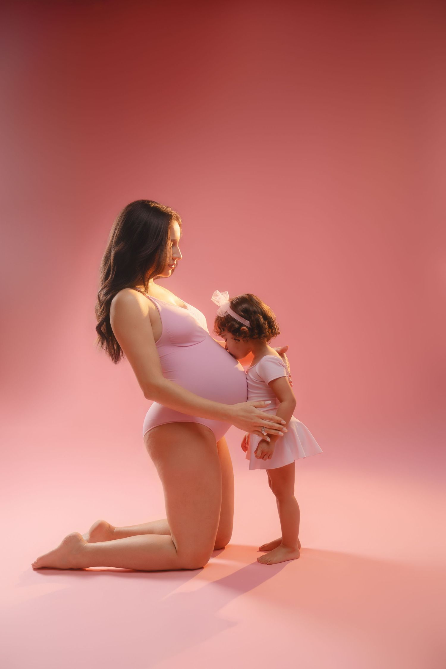Intimate maternity photoshoot shows parent and child bonding on pink studio floor.
