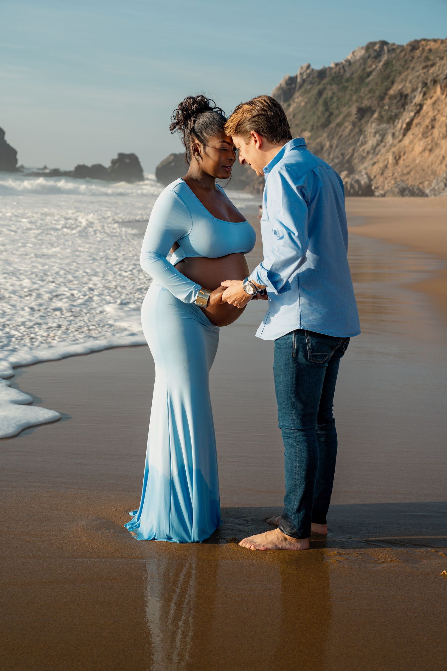 Pregnant couple holding hands on a sandy beach, woman in a blue maternity gown, man in a light blue shirt and jeans.