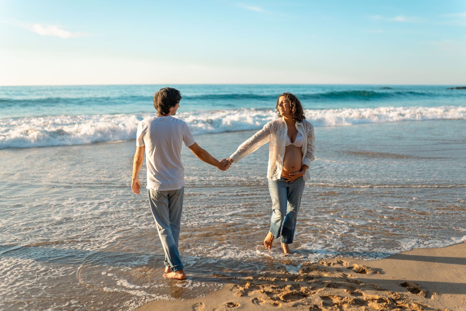 Couple walking hand in hand along ocean shore during sunset photo session.