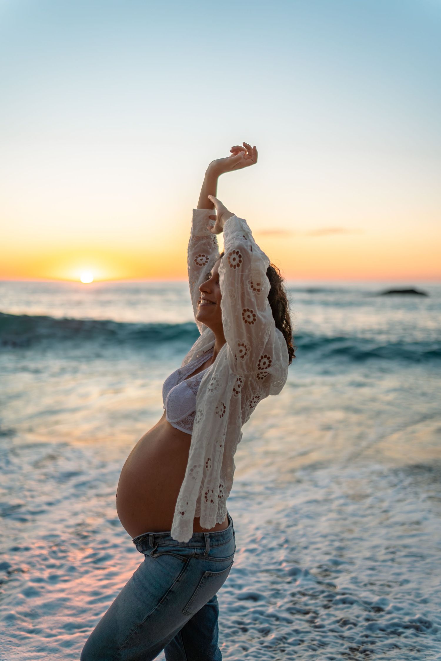 Maternity photoshoot at sunset on a beach with waves and golden light.