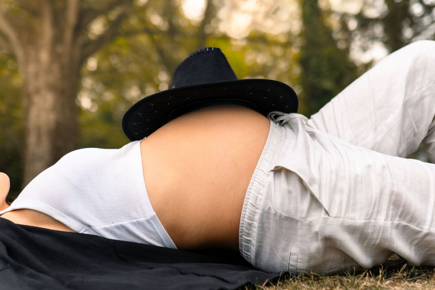 Grávida, deitada na relva numa posição relaxada durante um dia ensolarado ao ar livre, no campo, chapéu de cowboy na barriga