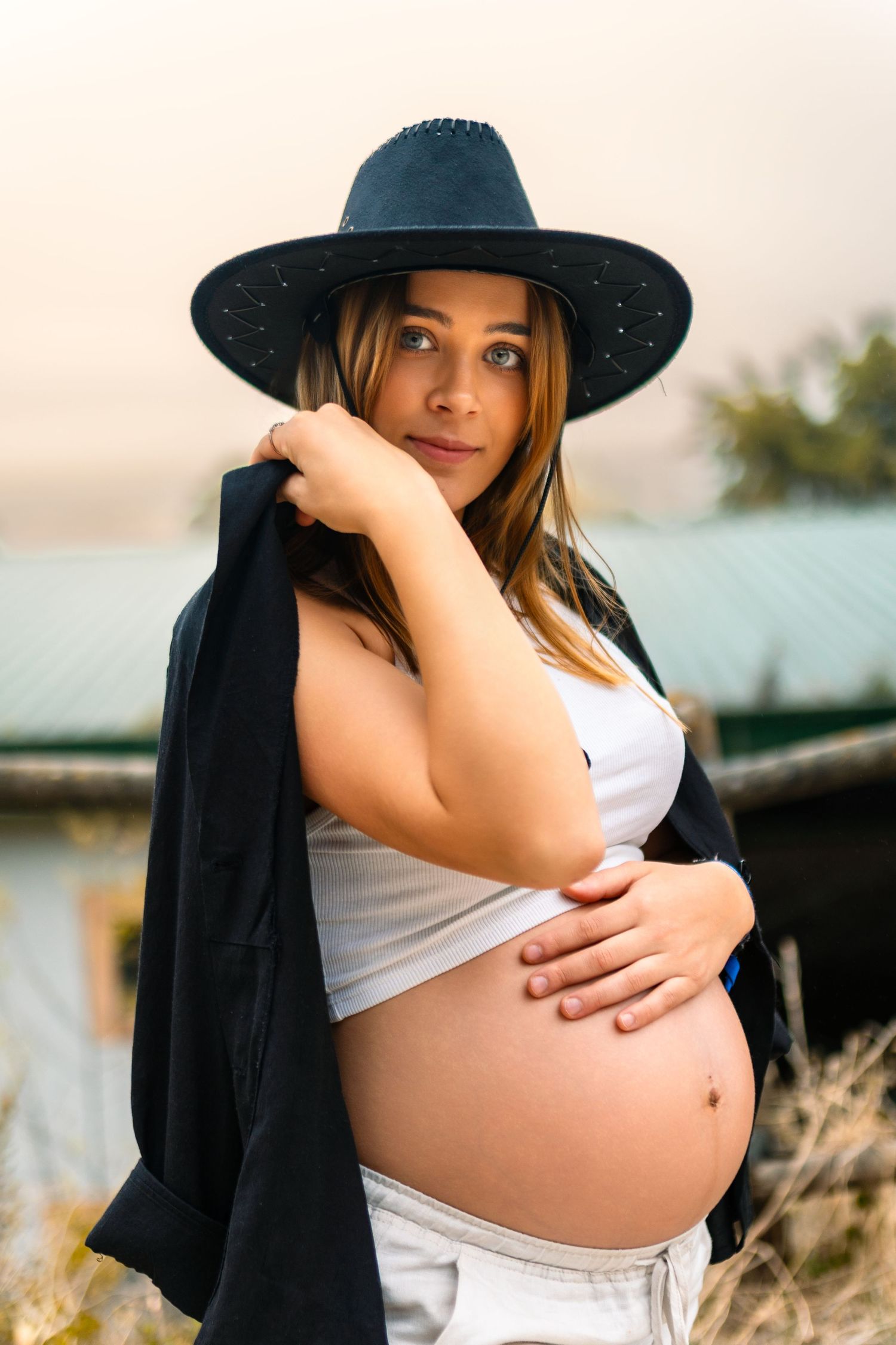 Uma futura mãe, grávida, de chapéu de cowboy preto, posa ao ar livre durante a golden hour, enquanto acaricia a barriga.