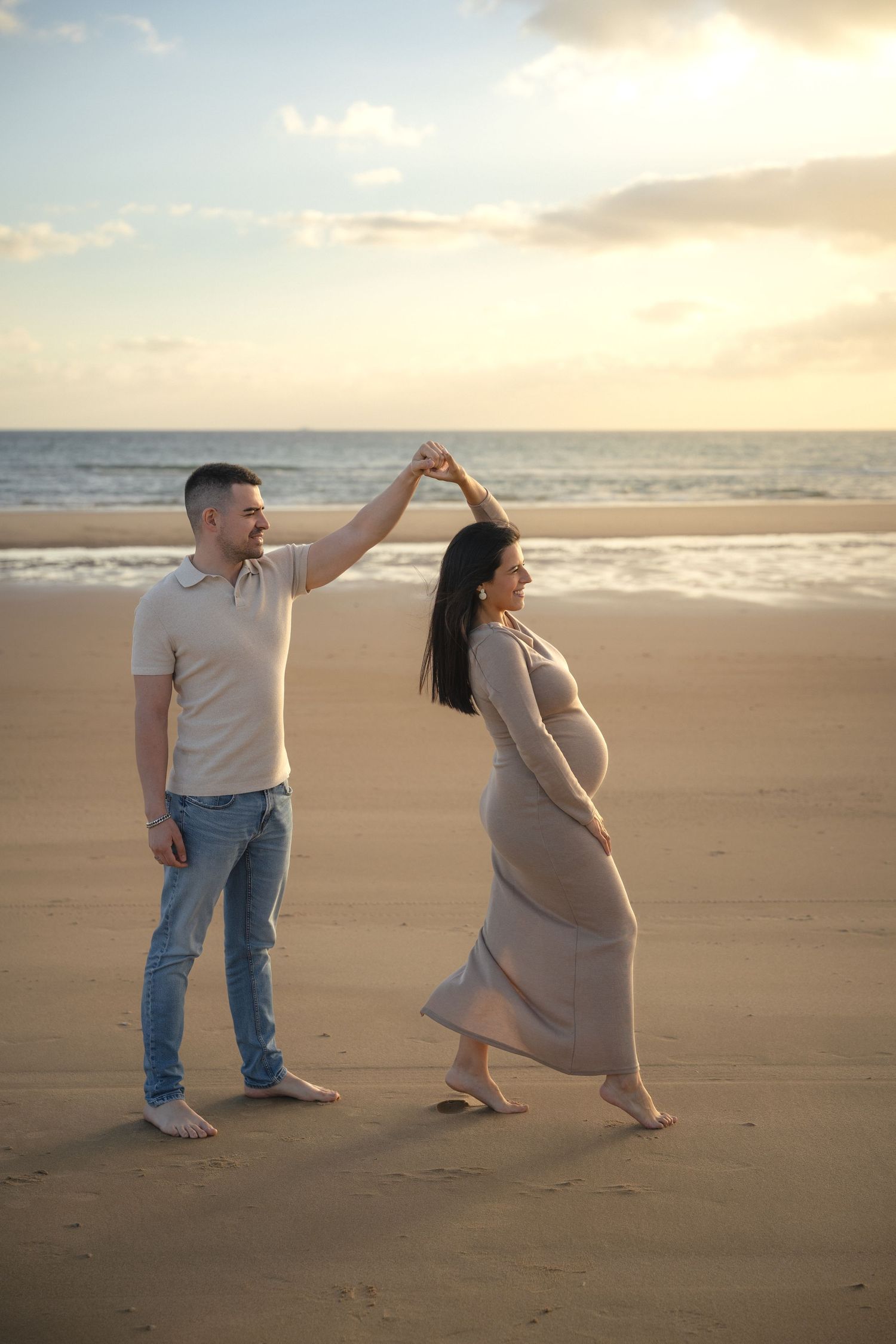Couple dancing on beach at sunset, pregnant woman in maxi dress twirled by partner in jeans near ocean waves.