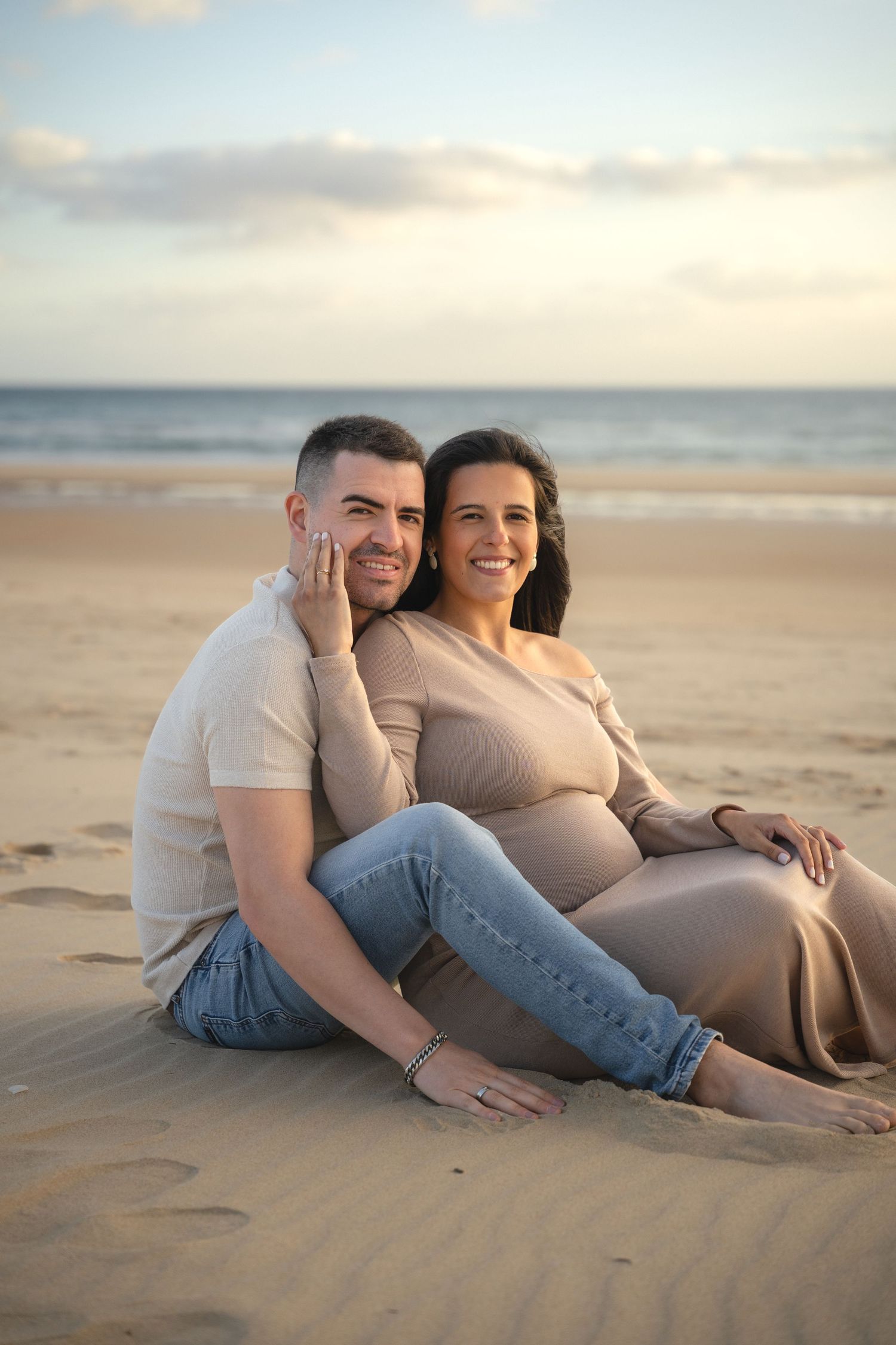 Couple sitting on sandy beach at sunset, pregnant woman in taupe dress leaning on partner in white shirt and jeans.