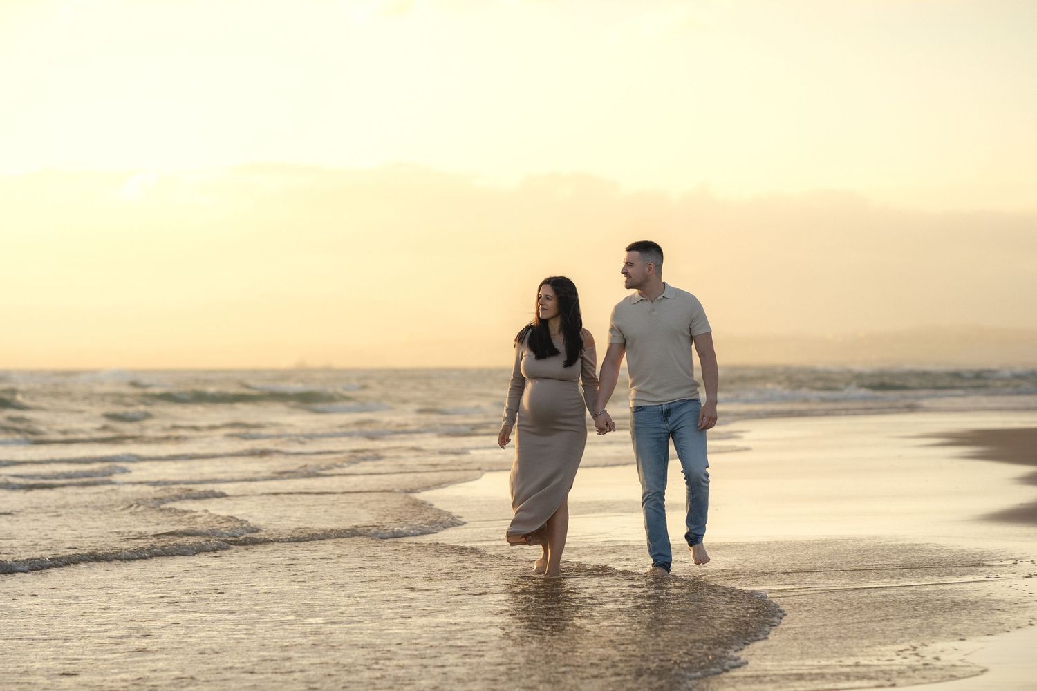 Couple walking hand in hand on a beach at sunset, waves gently washing over their feet.