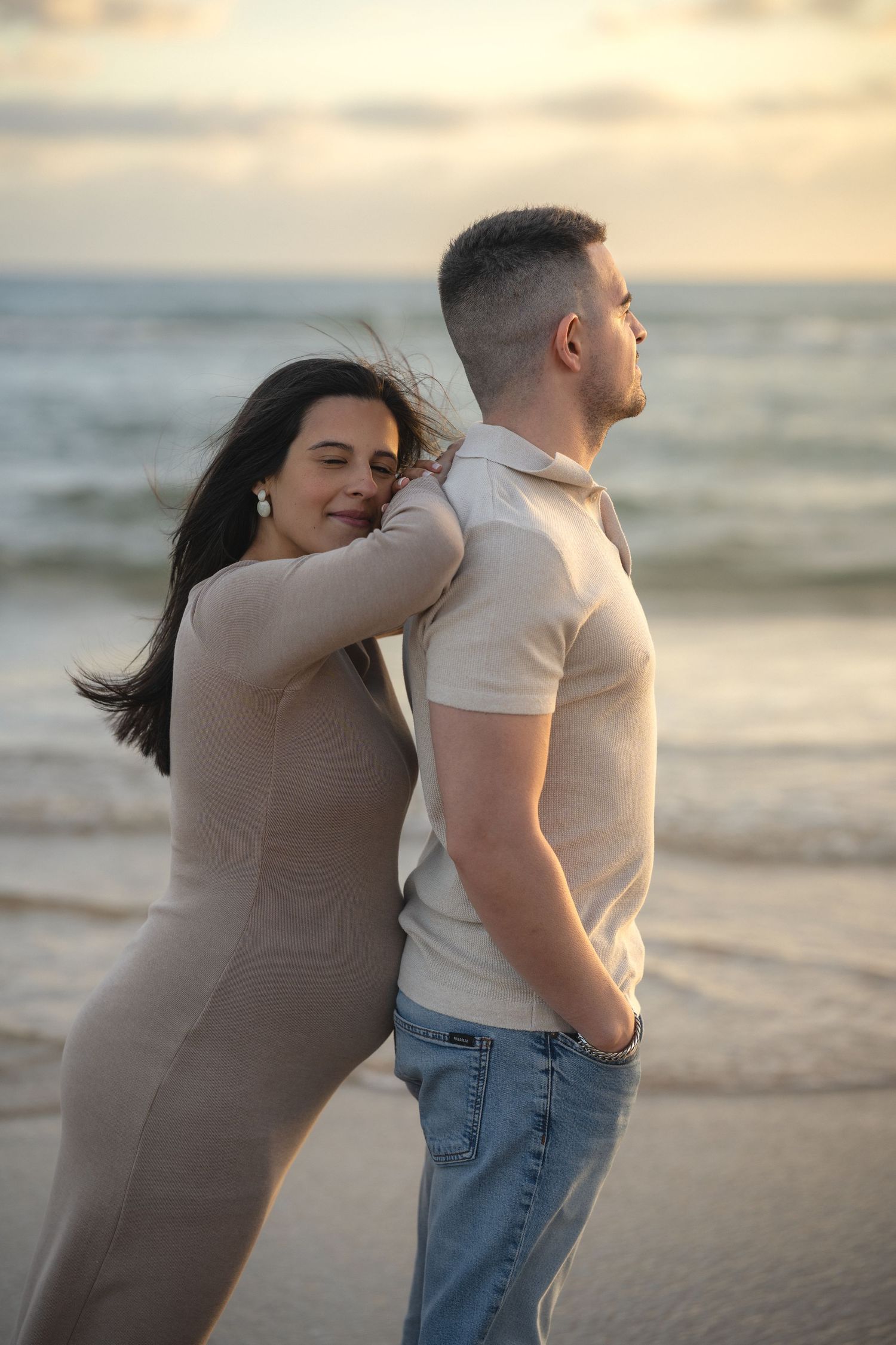 Couple embracing on a beach at sunset, woman in beige dress leaning on man in white polo and jeans.