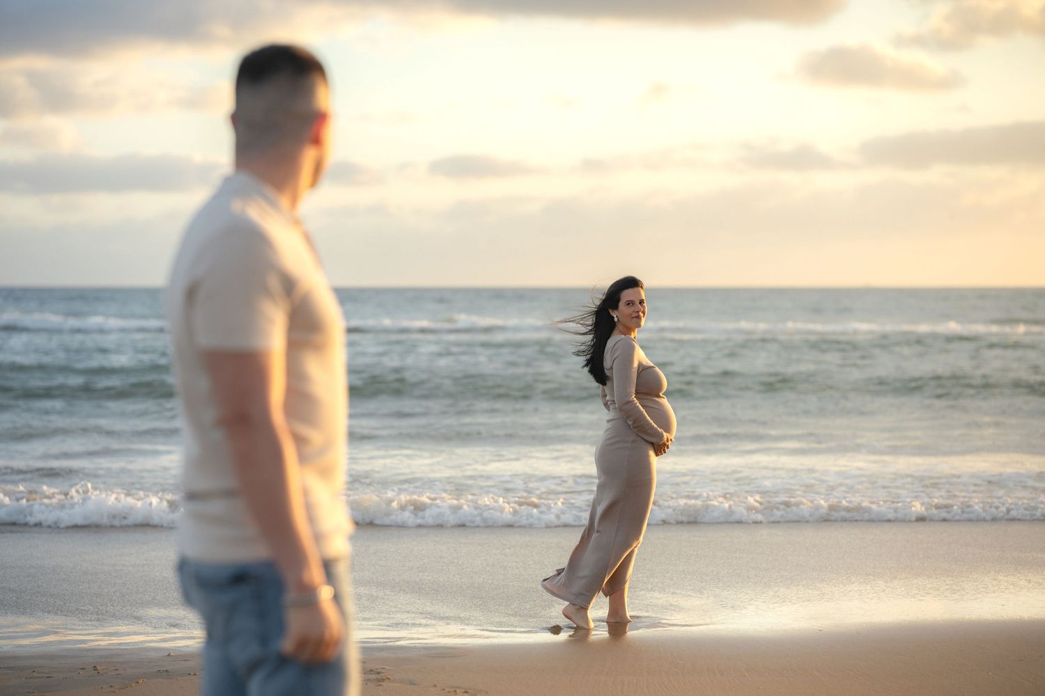 Pregnant woman in flowing dress poses on beach at sunset while man stands in foreground near ocean waves.