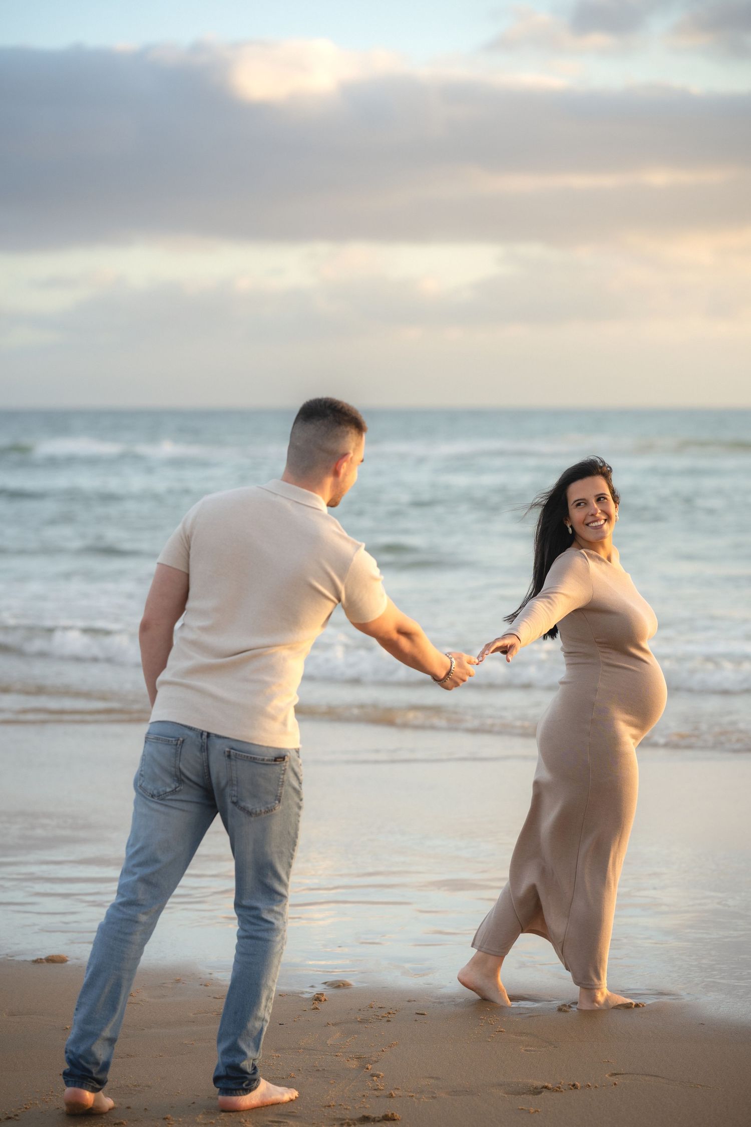 Pregnant couple holding hands and dancing on a sandy beach at sunset with ocean waves in the background.
