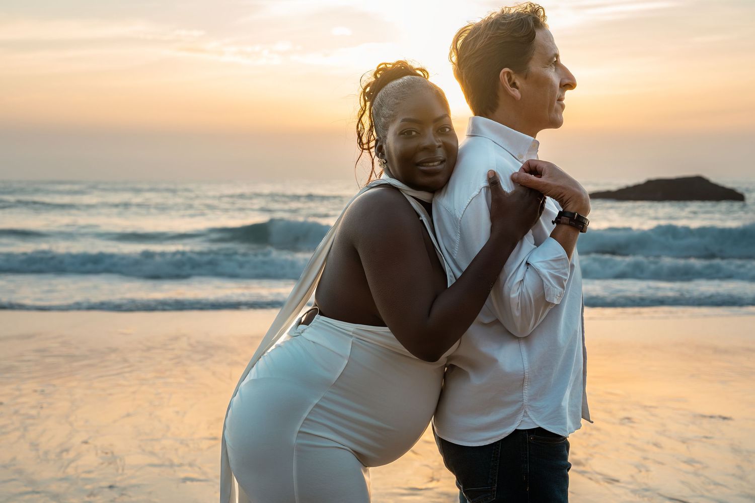Couple embracing on a beach at sunset, woman in white outfit leaning on man's back near ocean waves.