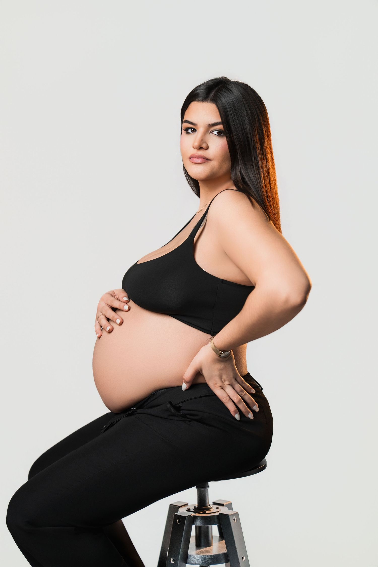 Elegant maternity photos showcase woman in black dress posing on modern stool.