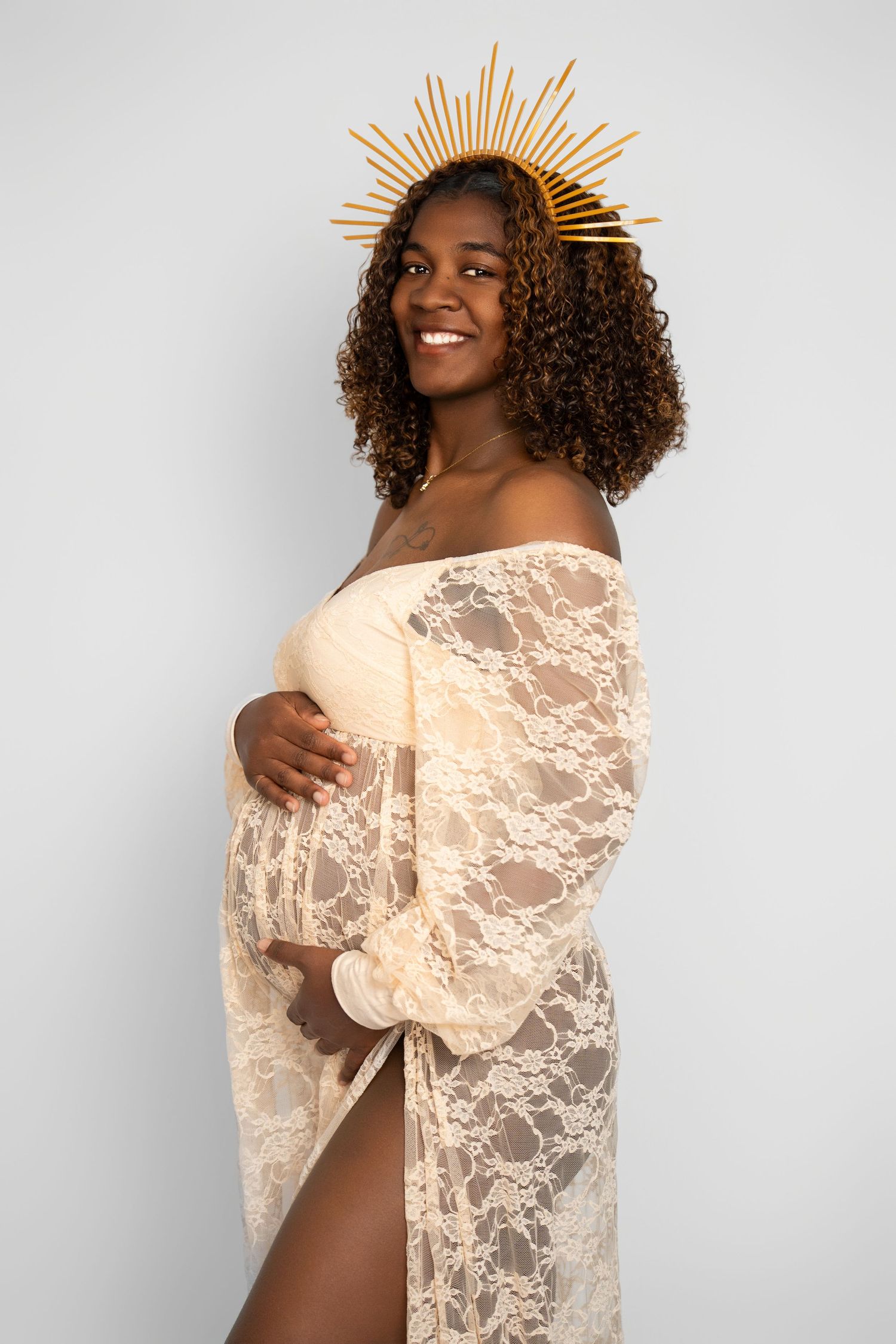 Maternity photo of a woman in a cream lace dress with curly hair wearing a golden sunburst crown against grey background.