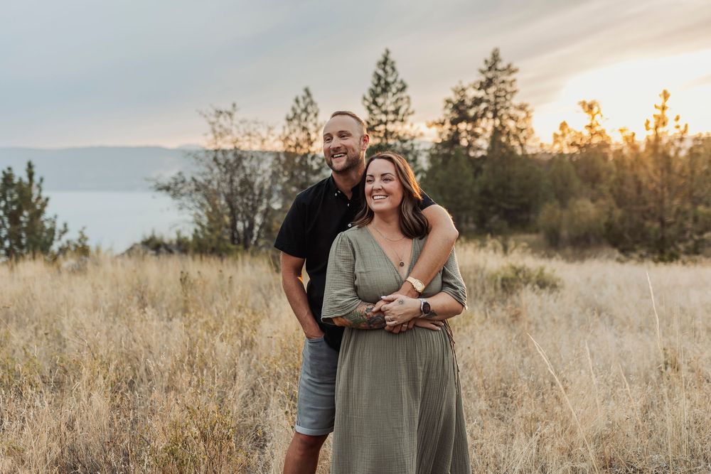 Couple embraces in golden field during sunset with mountains in background.