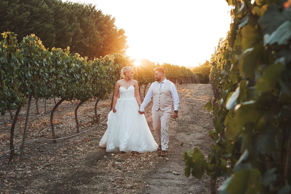 Bride and groom walking hand in hand through a sunlit vineyard at golden hour in a romantic outdoor wedding setting.