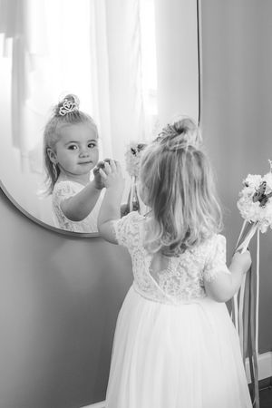 A young child in a white dress admires their reflection in a mirror.