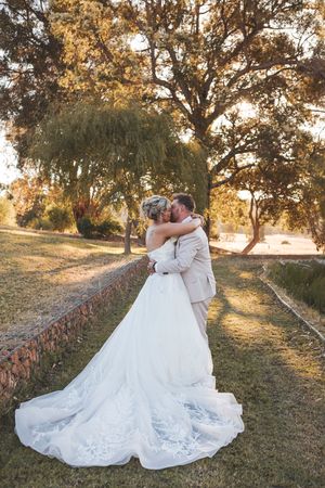 Bride and groom share a romantic kiss outdoors at golden hour, surrounded by lush trees, bride in elegant white gown with long train.