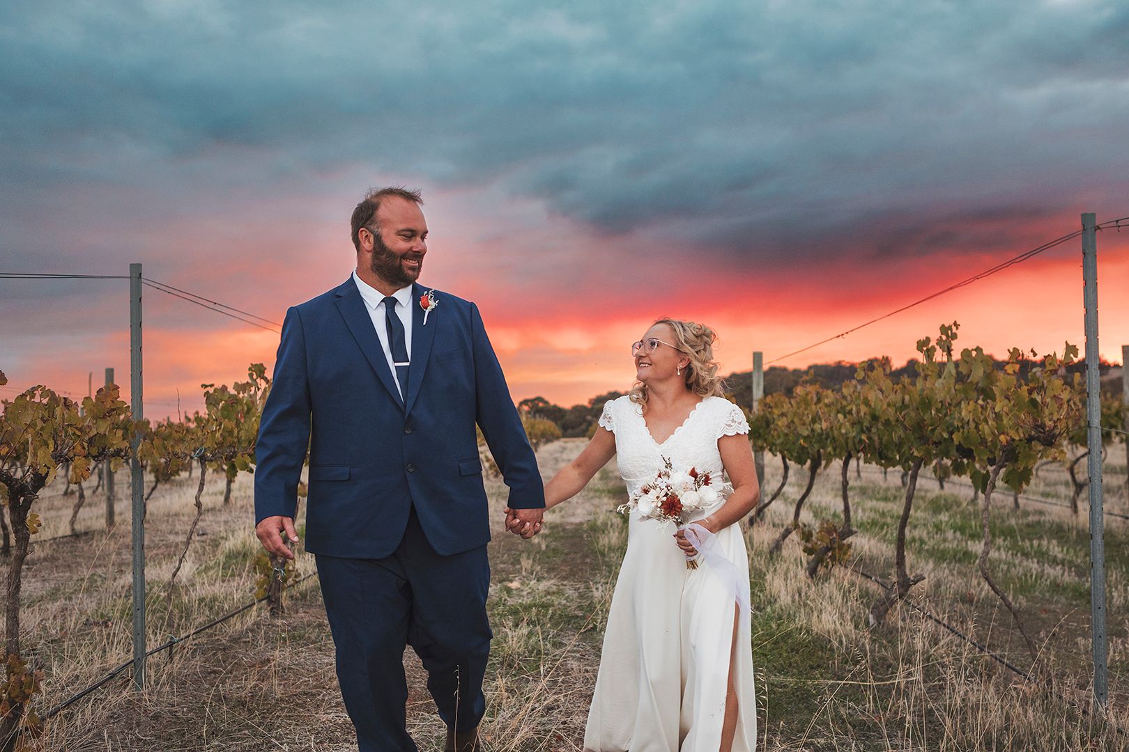 Newlyweds walk through vineyard rows against dramatic sunset sky.