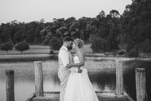 Black and white photo of a bride and groom sharing a kiss on a wooden dock by a serene lake surrounded by trees.