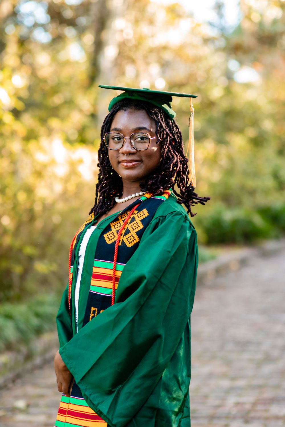 Graduate wearing a green commencement gown with traditional kente stole and decorative cords poses outdoors on a sunny day.