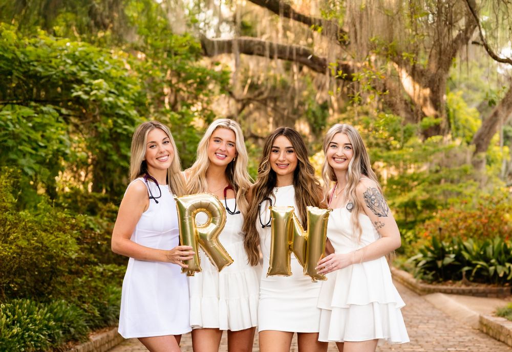 Four friends in white dresses pose together while holding gold letter balloons in a lush garden setting.