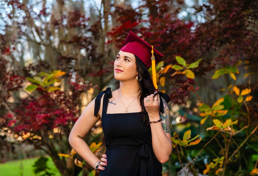 Someone wearing a burgundy graduate cap and black dress poses in front of vibrant autumn foliage in a garden setting.