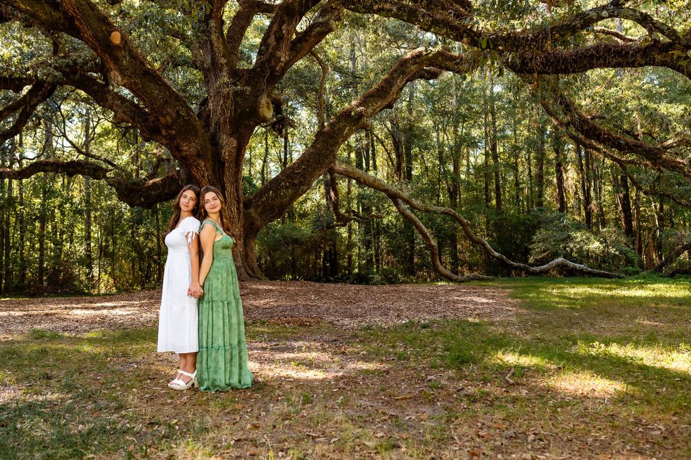 A couple in mint green dresses stands beneath majestic oak trees draped in Spanish moss at a scenic outdoor location.