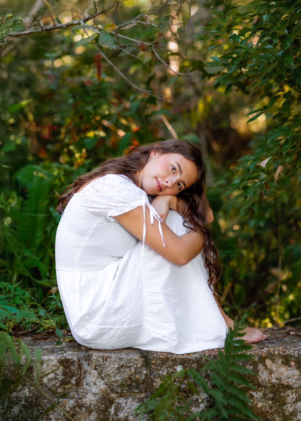 A young figure in a white dress reclines on a rustic stone wall surrounded by lush green foliage in natural sunlight.