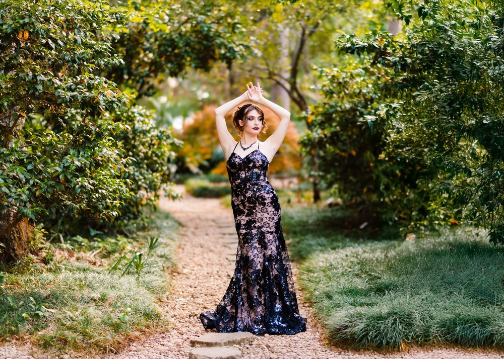 Woman in floral black gown poses gracefully on garden path surrounded by lush green trees.