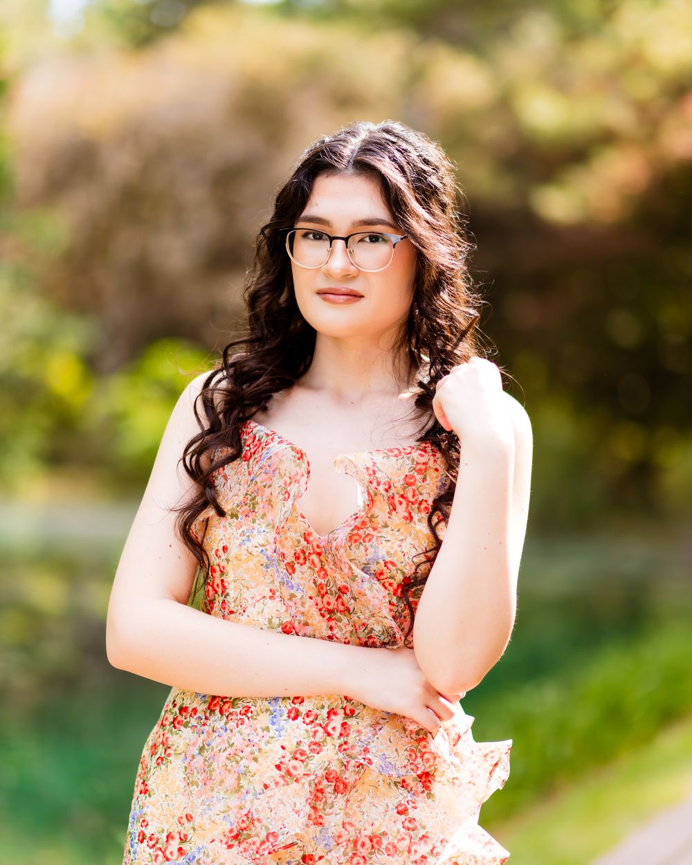 Young woman in floral dress and glasses posing outdoors in a lush green garden setting.