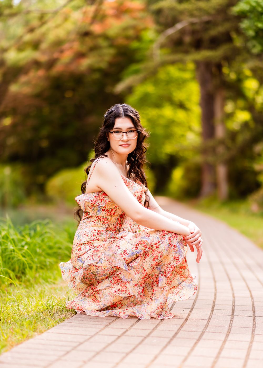 Young woman in floral maxi dress sitting gracefully on a wooden path surrounded by trees.