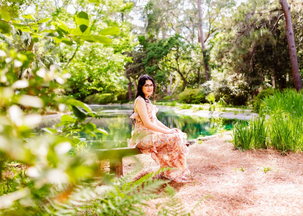 Woman in floral dress sitting by a serene garden pond surrounded by lush green trees and ferns.