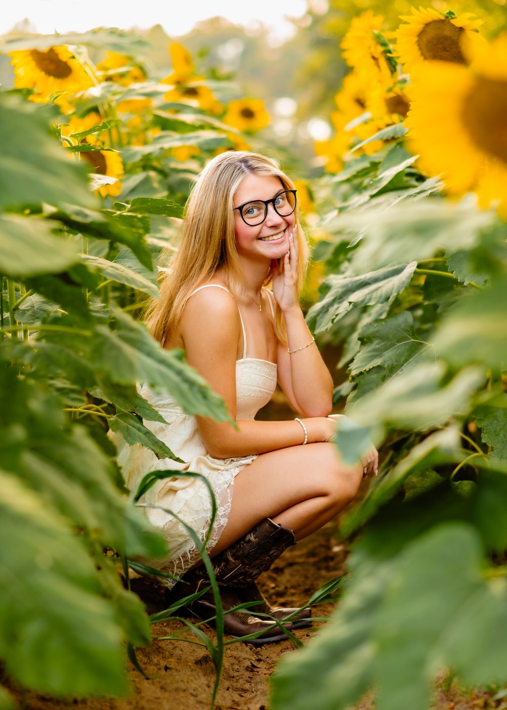 Young blonde woman with glasses crouching among tall sunflowers in a sunny field, smiling warmly in a white outfit.