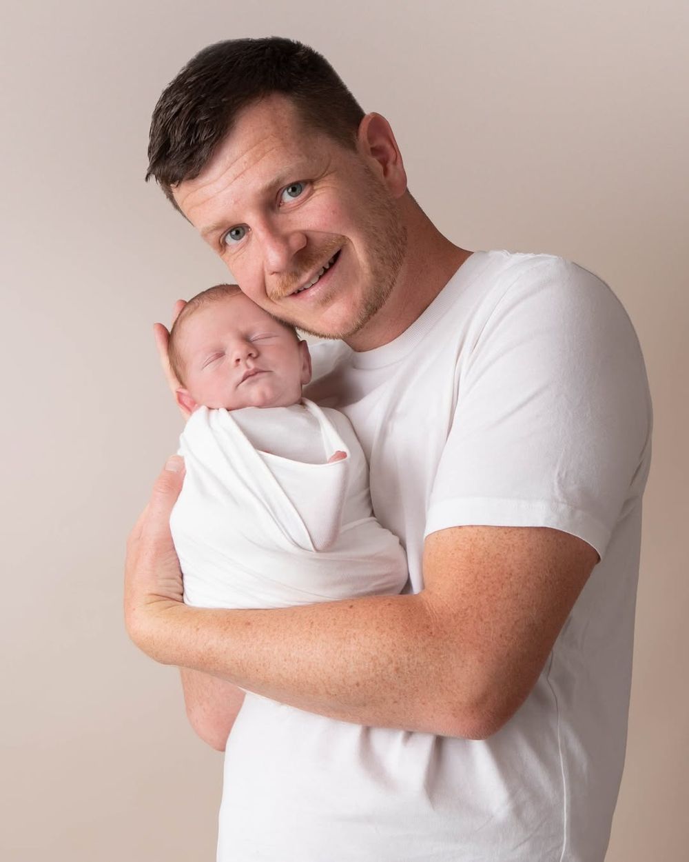 Smiling father in white t-shirt holds sleeping swaddled newborn baby in studio portrait.