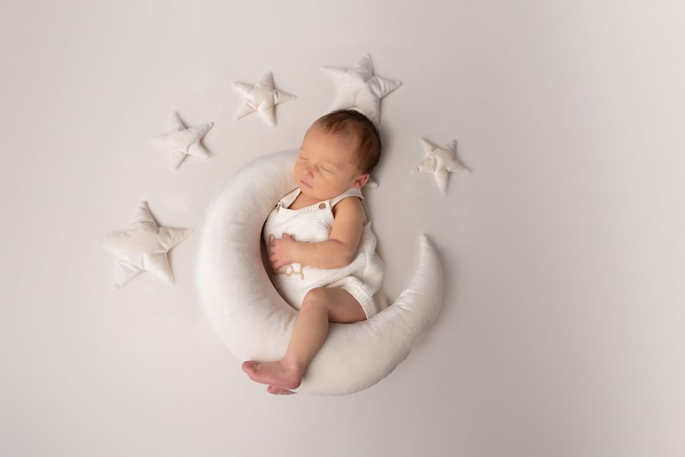 Newborn baby in white romper sleeping on a crescent moon pillow surrounded by star props on grey background.