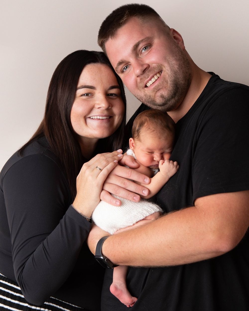 Smiling parents in black outfits proudly holding their sleeping newborn baby in a professional studio portrait.