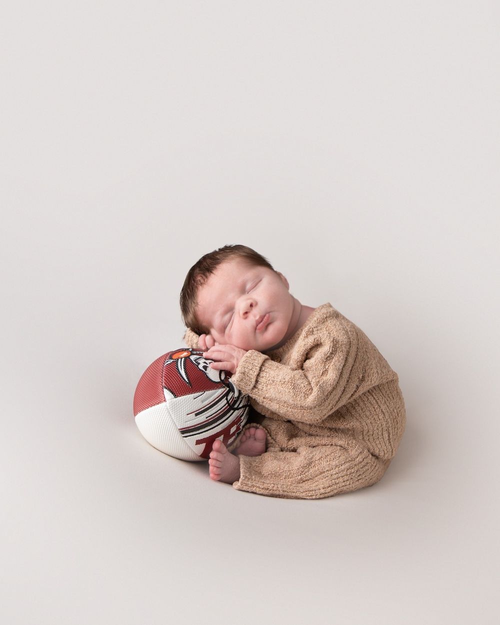 Sleeping newborn baby in brown knit outfit resting on a Tampa Bay Buccaneers football on white background.