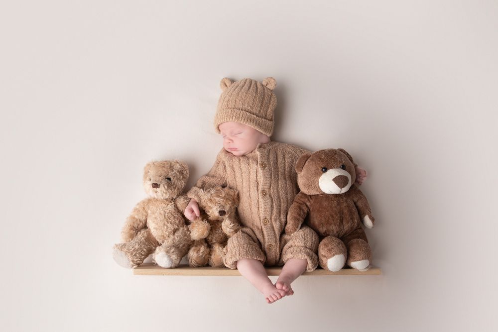 Adorable newborn baby in brown knit bear outfit sitting on a shelf surrounded by teddy bears.