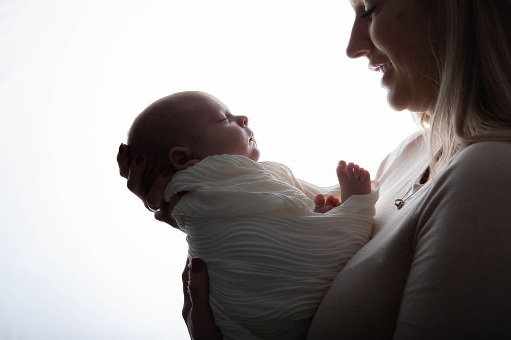 Silhouette of a smiling mother holding her alert newborn baby wrapped in white cloth.