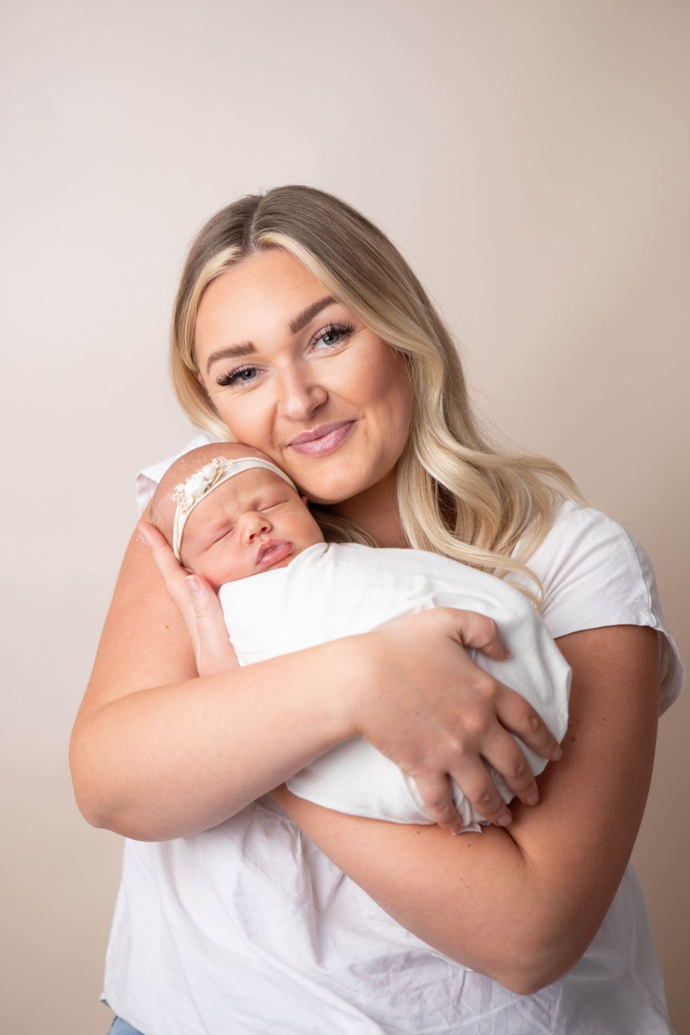Smiling blonde mother in white shirt tenderly holds her sleeping swaddled newborn baby girl.