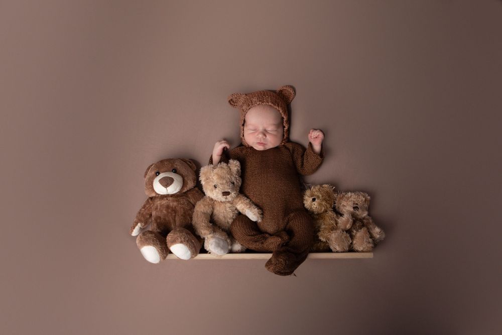 Sleeping newborn baby in brown bear outfit posed on shelf with three teddy bears.