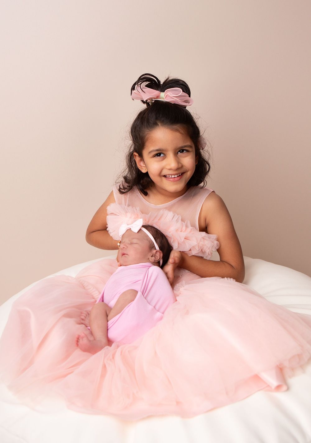 Smiling toddler girl in pink tulle dress holding sleeping newborn baby sister in a studio portrait.