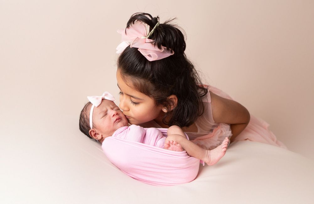 Young girl in pink dress lovingly kissing her swaddled newborn baby sibling in a studio photo.
