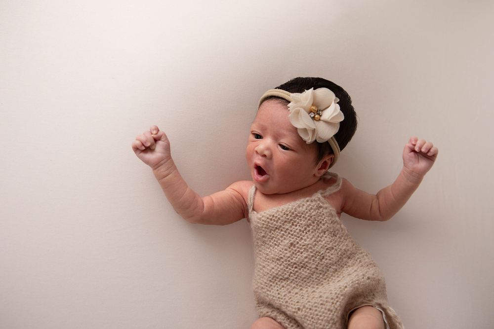 Adorable newborn baby girl in knit romper and floral headband stretches with arms raised wide.