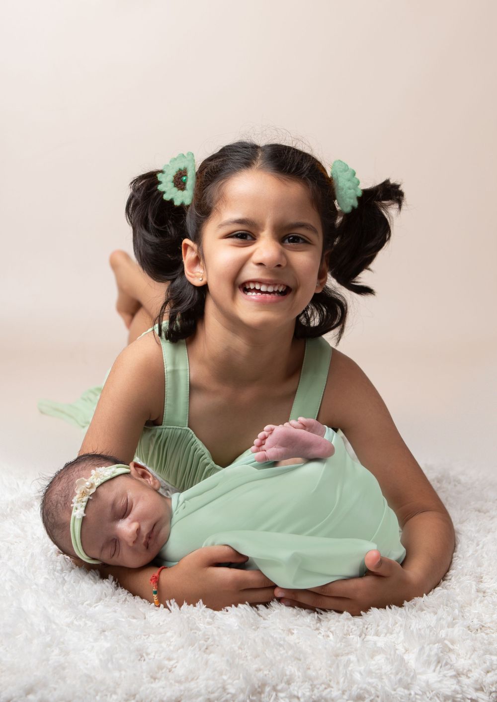 Joyful young girl in mint green dress smiling while holding her swaddled newborn baby in studio portrait.