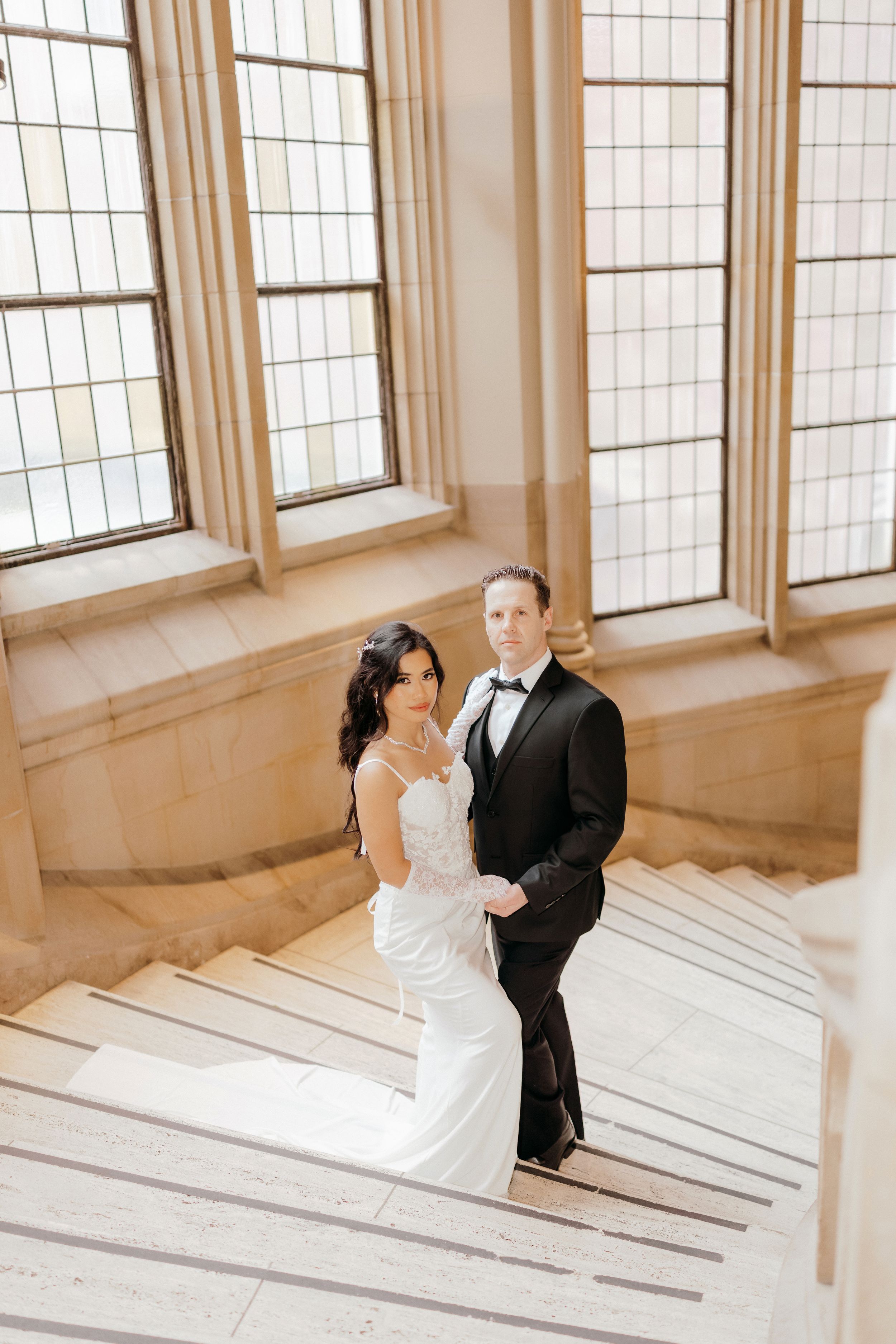 A romantic wedding couple poses in an elegant white room with large windows creating a bright, airy atmosphere.